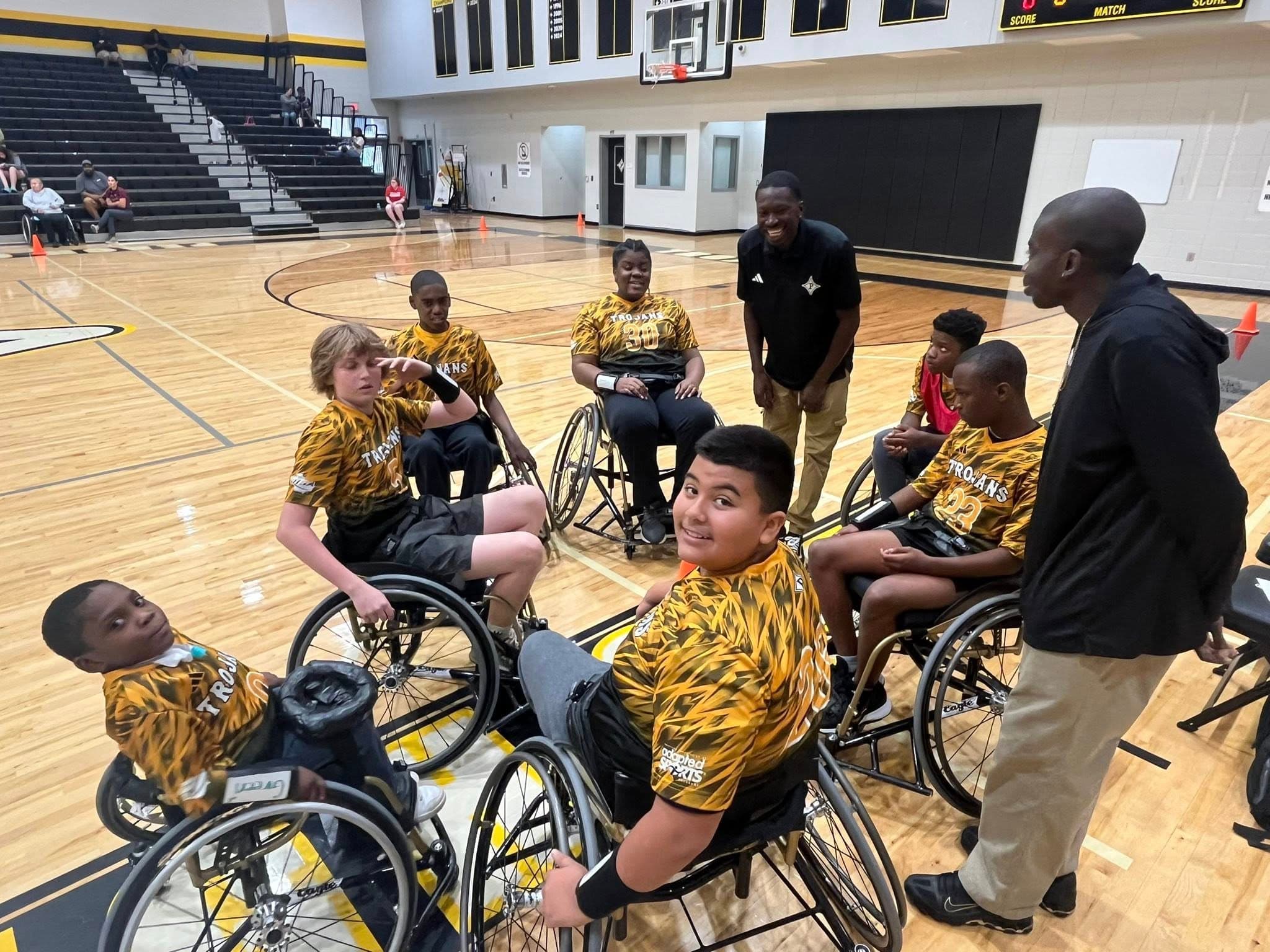 A wheelchair basketball team with players and coaches in yellow jerseys gather on an indoor court for a discussion.